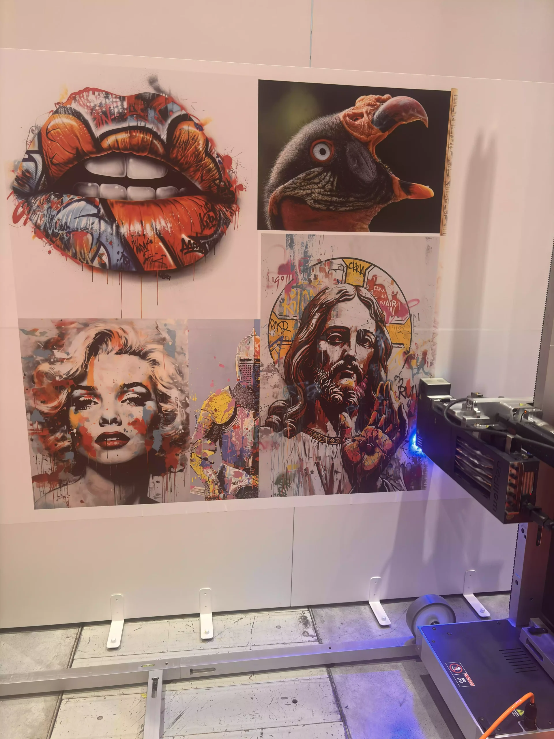 Pionowa drukarka ścienna UV w trakcie pracy nanosząca wydruk artystyczny obok gotowej galerii portretów (Marilyn Monroe, Jezus, usta graffiti, ptak)